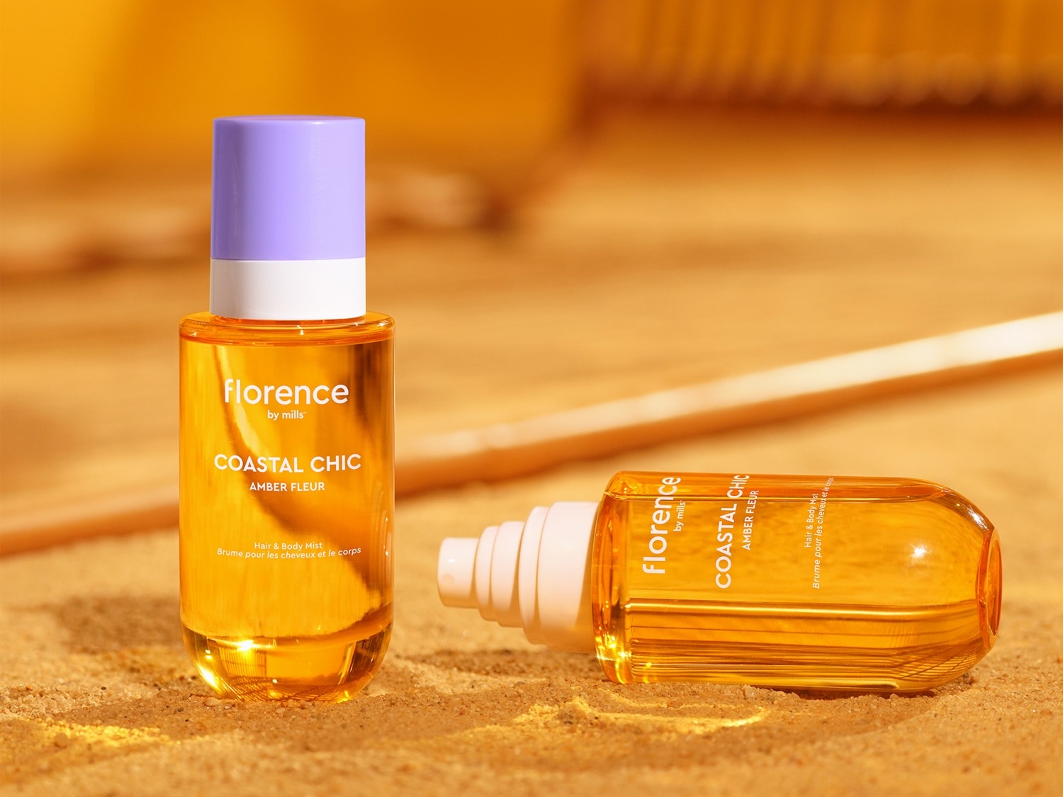 Two Florence by Mills Coastal Chic Amber Fleur Hair & Body Mist bottles, one standing upright with a lavender cap and the other lying on its side, both filled with a golden liquid, are displayed on a sandy surface.