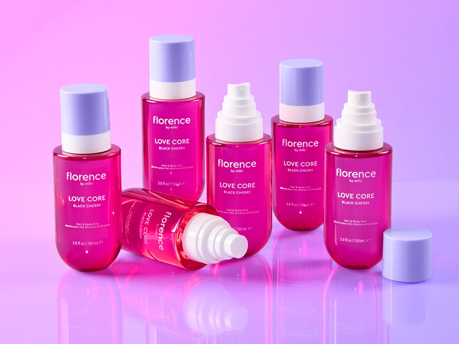 Six Florence by Mills Love Core Black Cherry Hair & Body Mist bottles, five standing upright and one lying on its side, are displayed on a vibrant purple and pink gradient background, each featuring a bright pink liquid and a light purple cap, with some bottles having white spray nozzles visible.