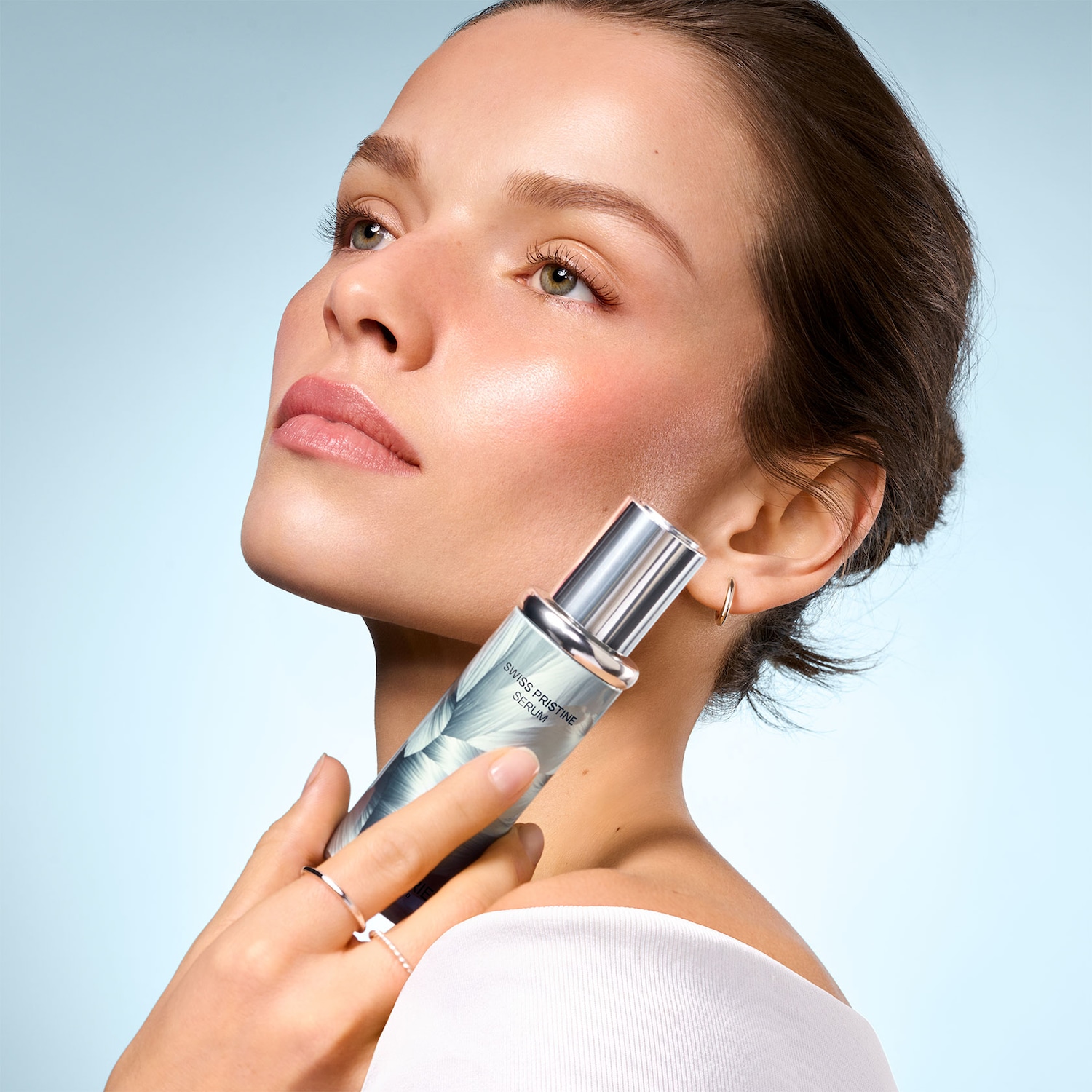 Eine Frau mit makelloser Haut hält ein silbernes Serumfläschchen mit der Aufschrift "SWISS PRISTINE SERUM" an ihren Hals, ihr Blick ist nach oben gerichtet.
