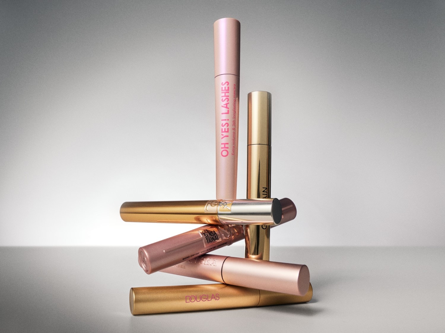 Een stapel van zes mascara's in goud- en roségoudtinten, met de roze "OH YES! LASHES" mascara prominent bovenaan, klaar om je wimpers te transformeren.