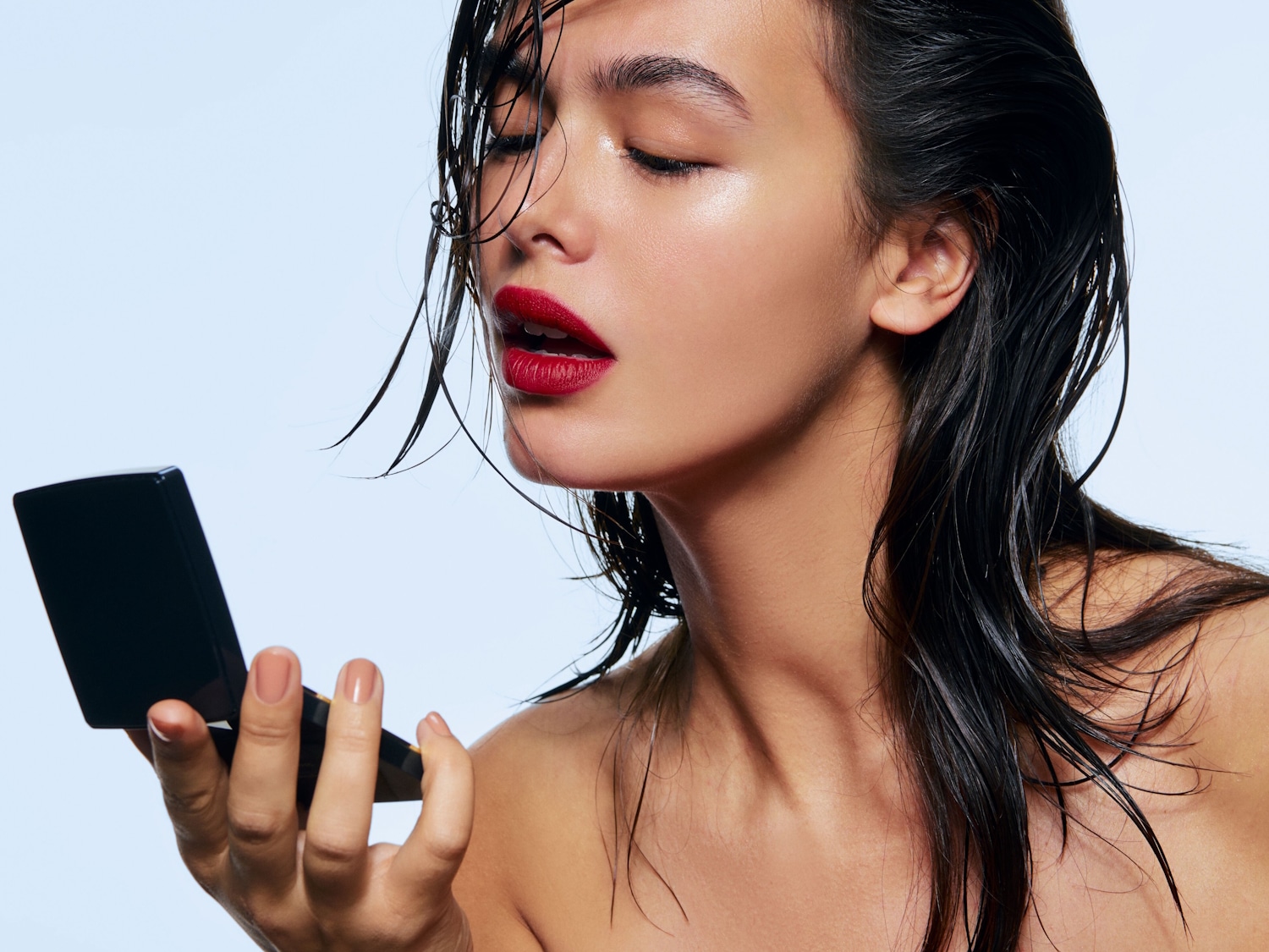 Een vrouw met nat, donker haar en glanzende huid, met gesloten ogen en felrode lippen, kijkt in een compact zwart spiegeltje.