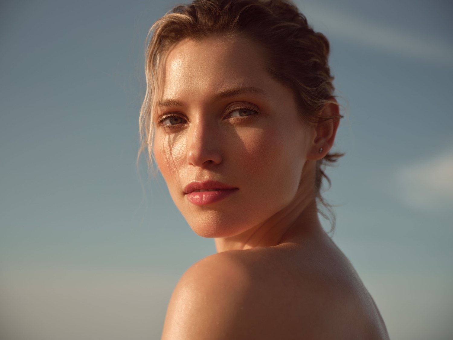 Een vrouw met een stralende huid en natuurlijke make-up kijkt over haar schouder, met de zon die haar gezicht en schouders verlicht tegen een helderblauwe lucht.