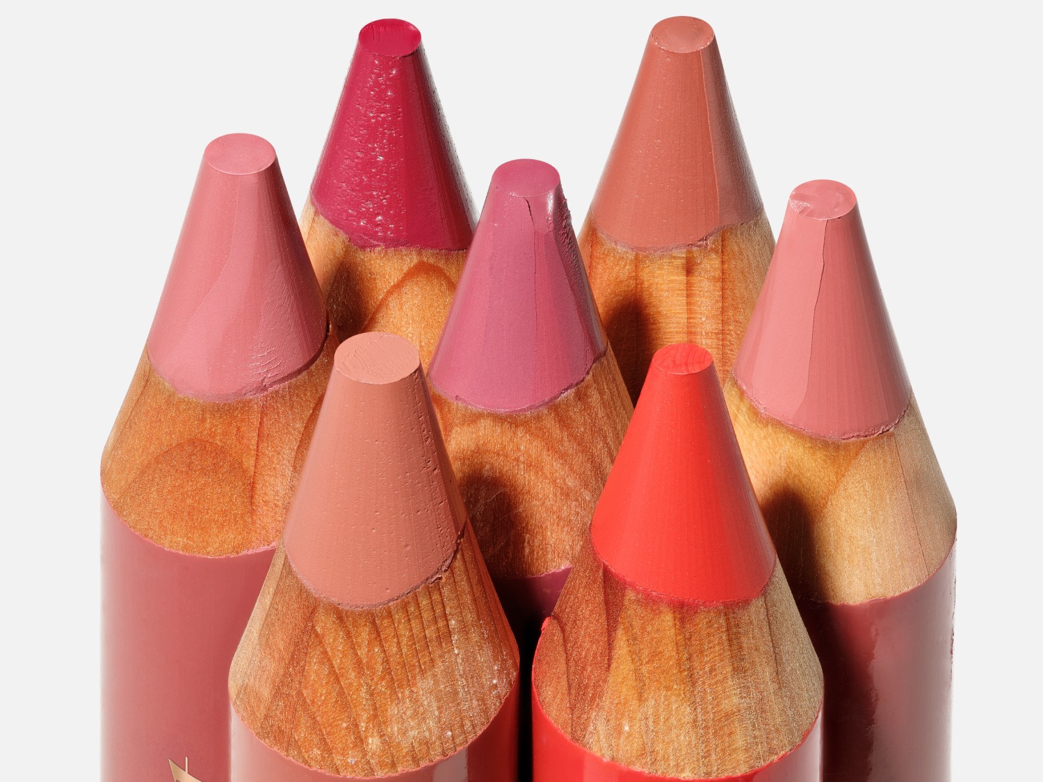 Een close-up van zeven lippenstiftpotloden in verschillende roze en rode tinten, met houten omhulsels en puntige, romige stiften.