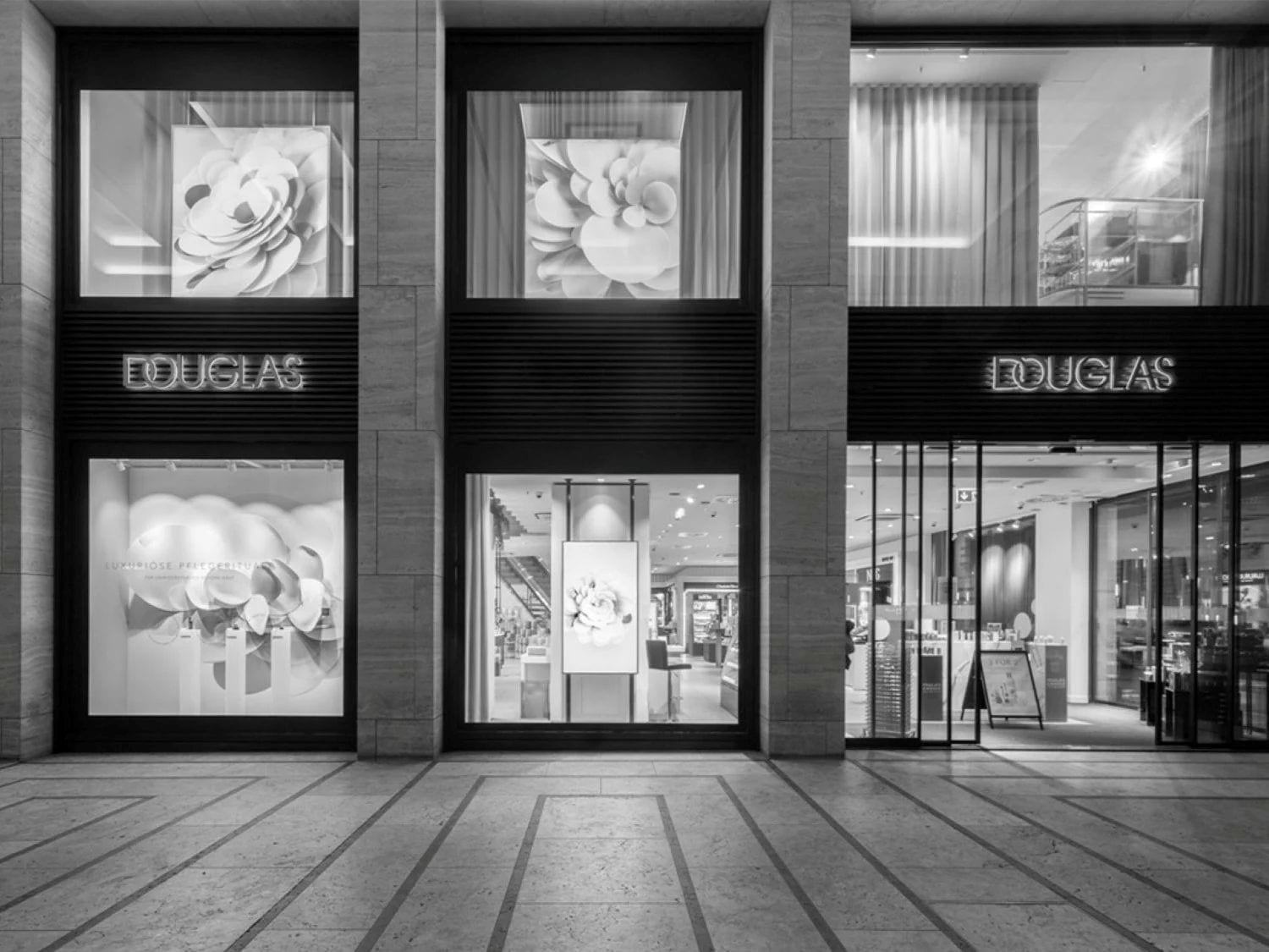 La facciata del negozio Douglas presenta vetrine con grandi immagini di fiori bianchi e il logo Douglas illuminato, invitando i clienti a scoprire le lussuose collezioni di prodotti di bellezza.