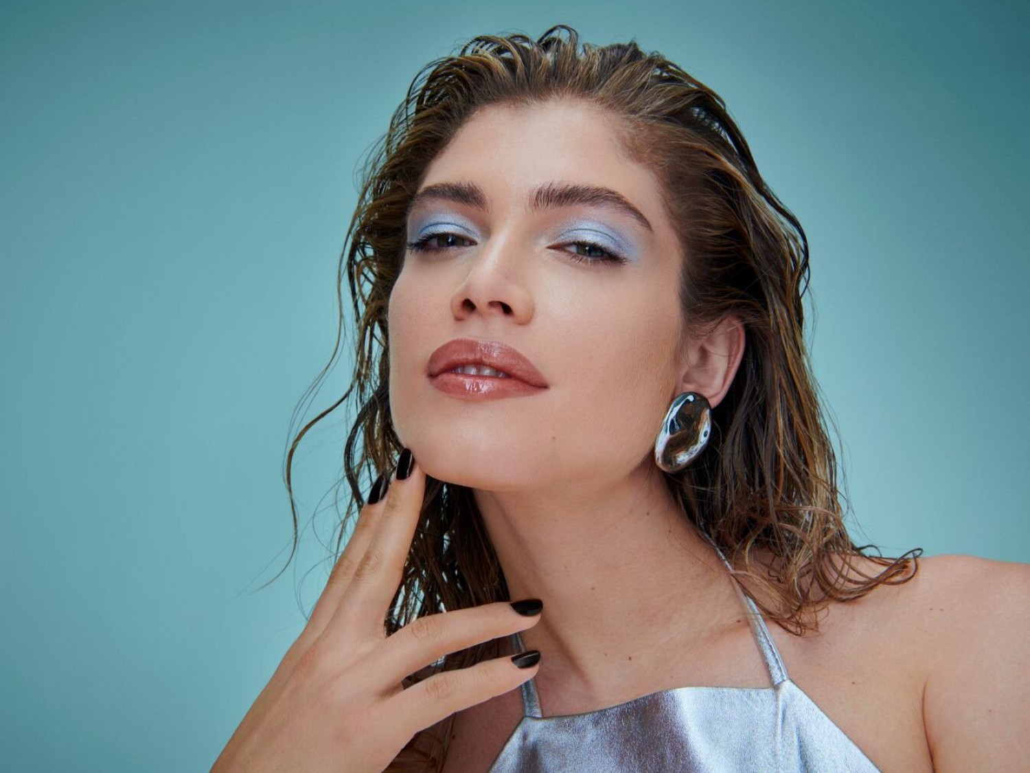 Una donna con capelli bagnati e lucidi, trucco azzurro sugli occhi e labbra lucide, indossa un top argentato e orecchini a bottone argentati, con unghie nere, su sfondo azzurro.