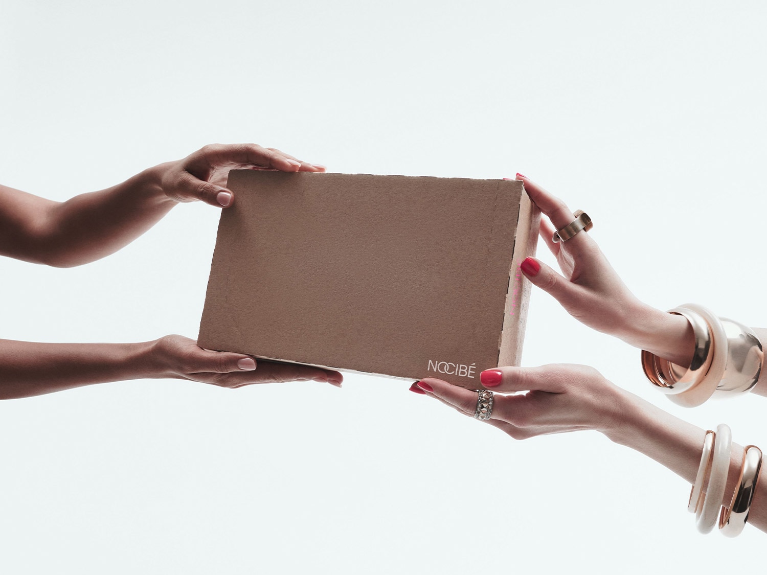 Deux paires de mains échangent une boîte en carton marron clair avec le logo Nocibé, les mains de droite portent des bagues et des bracelets dorés et ont les ongles peints en rouge.