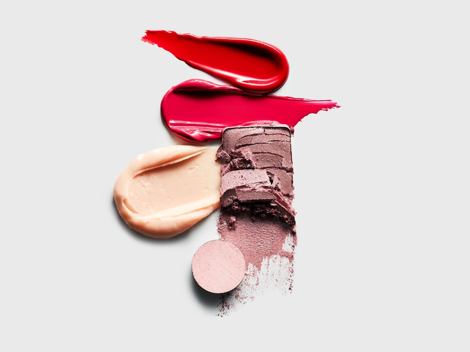 Een close-up van verschillende make-upstalen, waaronder twee rode lipgloss-vegen, een beige crème-achtige substantie en twee tinten paarse oogschaduw, één in poedervorm en één geperst.