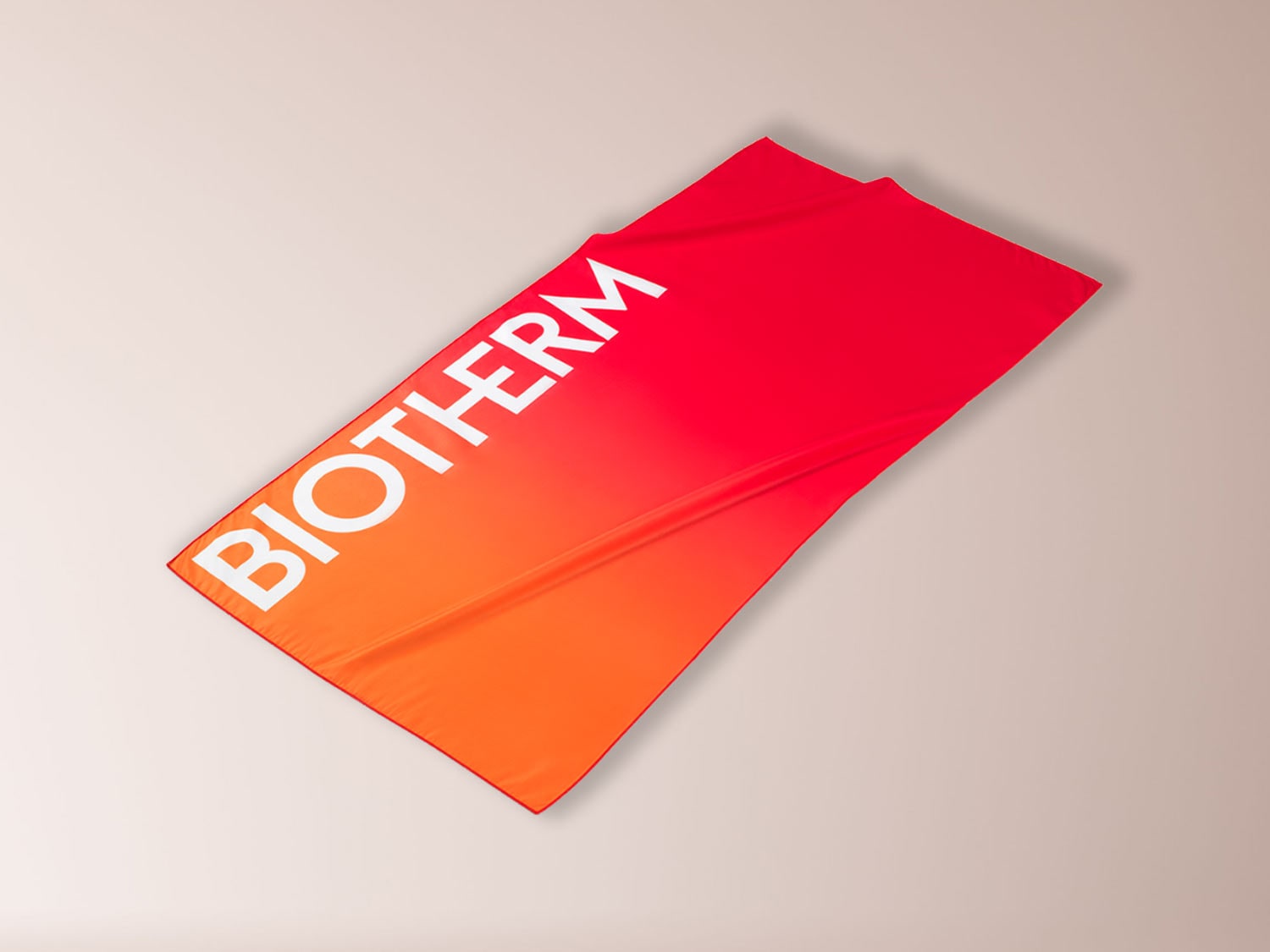 Ein rotes und orangefarbenes Handtuch mit dem weißen Schriftzug "BIOTHERM" ist diagonal auf einem hellbraunen Hintergrund platziert.