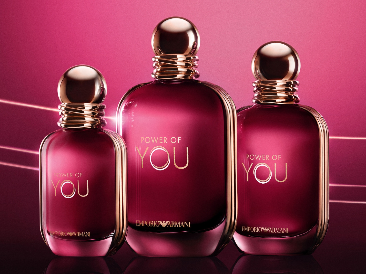 Drie flessen Emporio Armani "Power of You" parfum, variërend in grootte, met een dieprode vloeistof en gouden accenten, staan tegen een levendige roze achtergrond.
