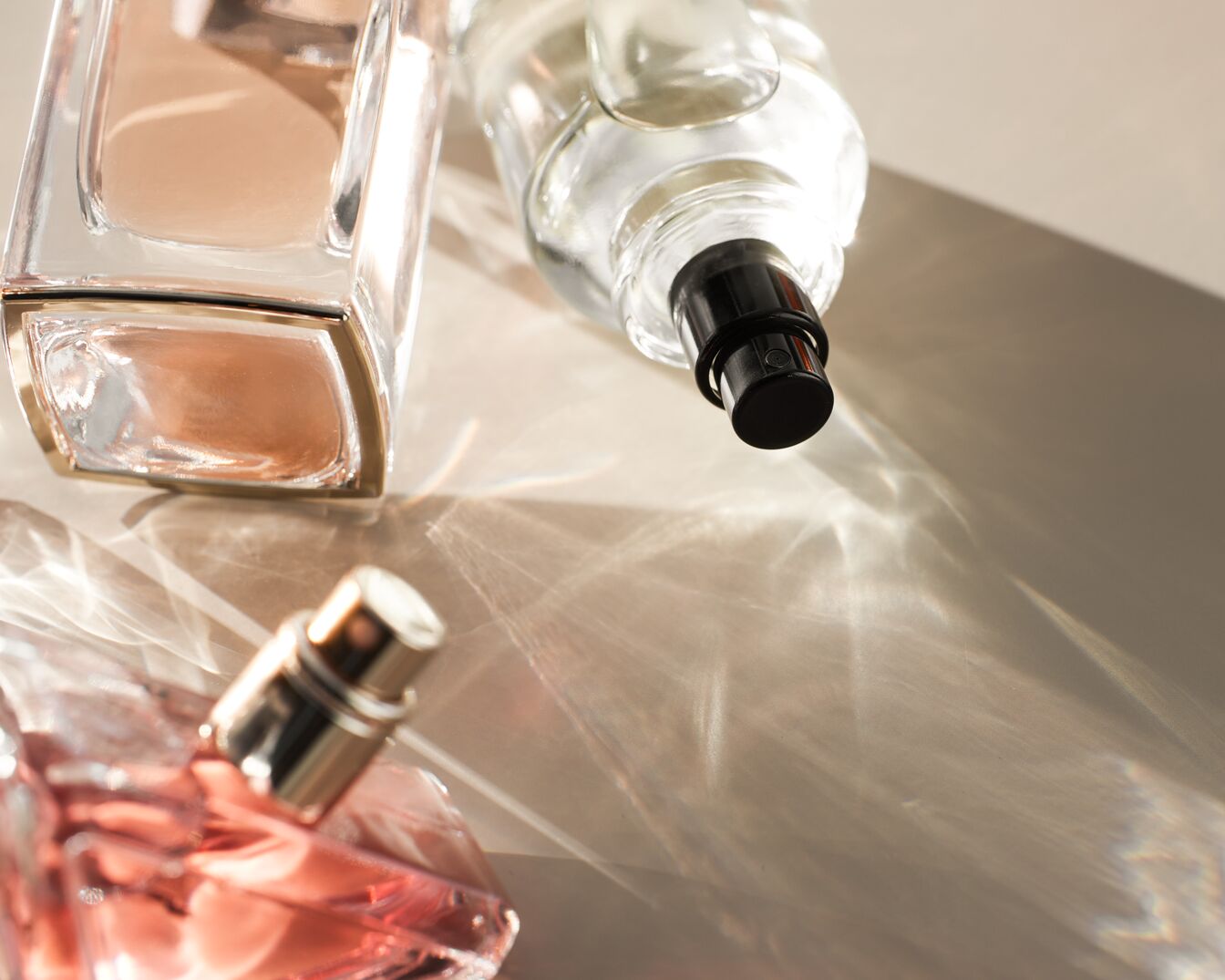 Trois flacons de parfum en verre, un avec un liquide beige, un autre avec un liquide rose et un troisième transparent, sont posés sur une surface claire avec des reflets lumineux.