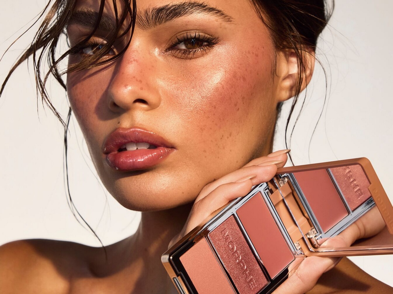 Une femme à la peau bronzée et aux cheveux mouillés tient une palette de maquillage Morphe avec des fards à joues et des surligneurs dans des tons pêche et bronze, mettant en valeur un look frais et éclatant.
