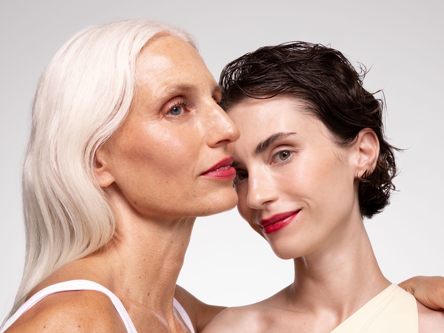Twee vrouwen, de een met wit haar en de ander met donkerbruin haar, leunen tegen elkaar aan, beide met een stralende huid en rode lippenstift.