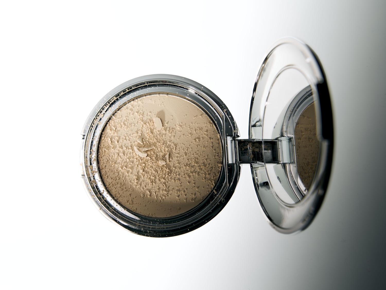 Een open compactpoederdoosje met een zilveren rand en een spiegel in het deksel, gevuld met lichtbeige poeder dat deels verkruimeld is.