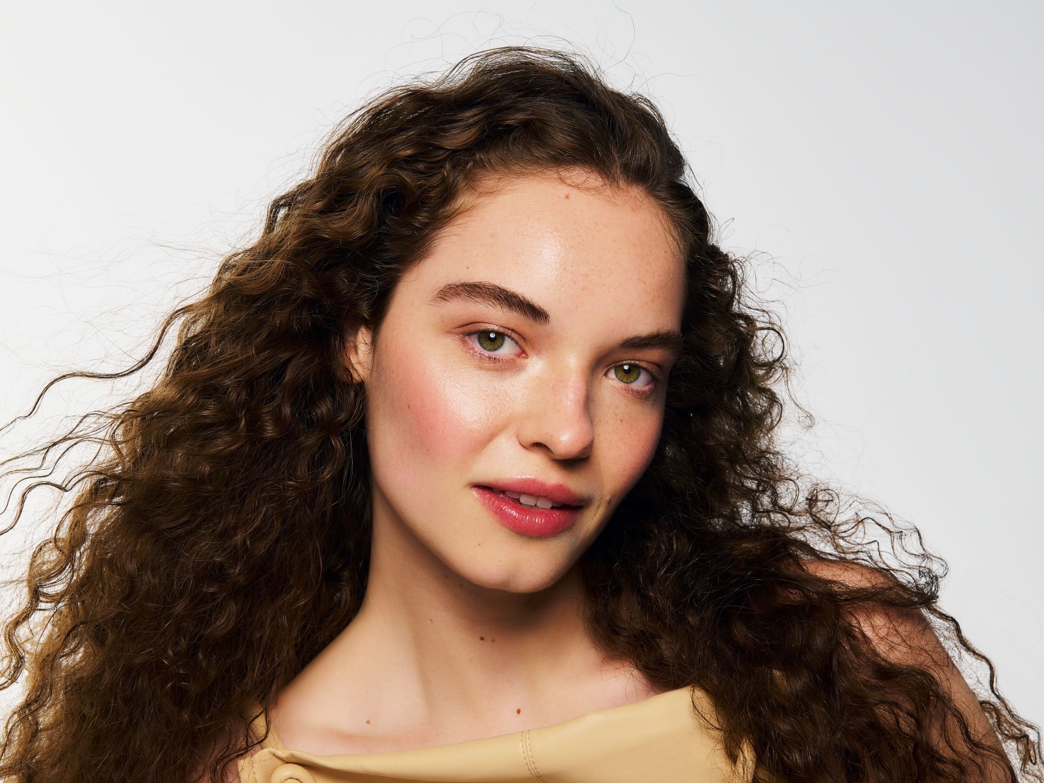 Een jonge vrouw met een lichte huid, sproeten en lang, donkerbruin krullend haar kijkt recht in de camera, haar gezicht is lichtroze en haar lippen zijn glanzend roze.