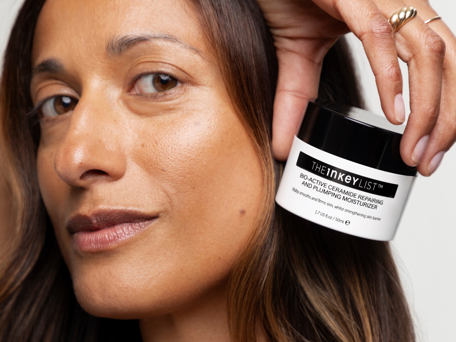 Eine Frau mit braunen Haaren und gebräunter Haut hält einen weißen Tiegel der Marke The Inkey List mit schwarzem Deckel und der Aufschrift Bio-Active Ceramide Repairing and Plumping Moisturizer neben ihrem Gesicht.