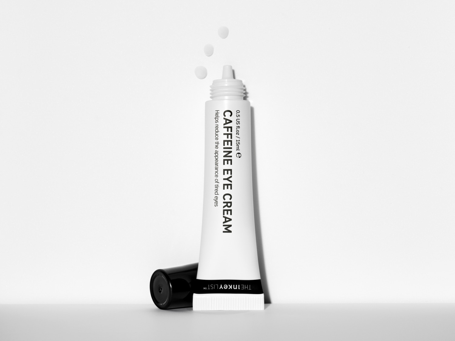 Eine weiße Tube der The Inkey List Caffeine Eye Cream mit schwarzer Schrift und abgenommener schwarzer Kappe, aus der drei Tropfen der Creme austreten, liegt auf weißem Untergrund.
