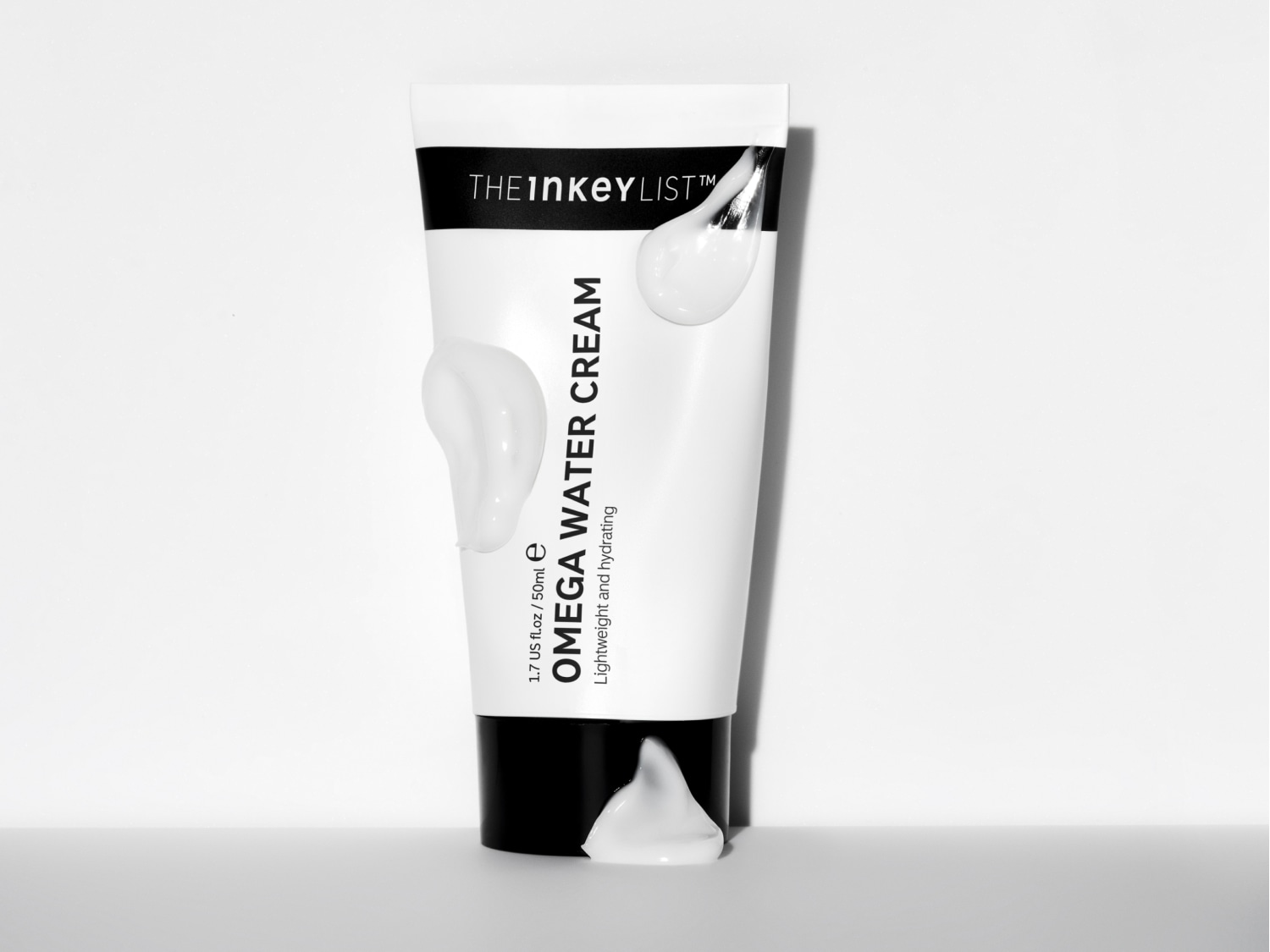 Eine weiße Tube der Inkey List Omega Water Cream mit schwarzer Schrift und einem schwarzen Streifen ist mit mehreren, weißen Klecksen der Creme versehen.
