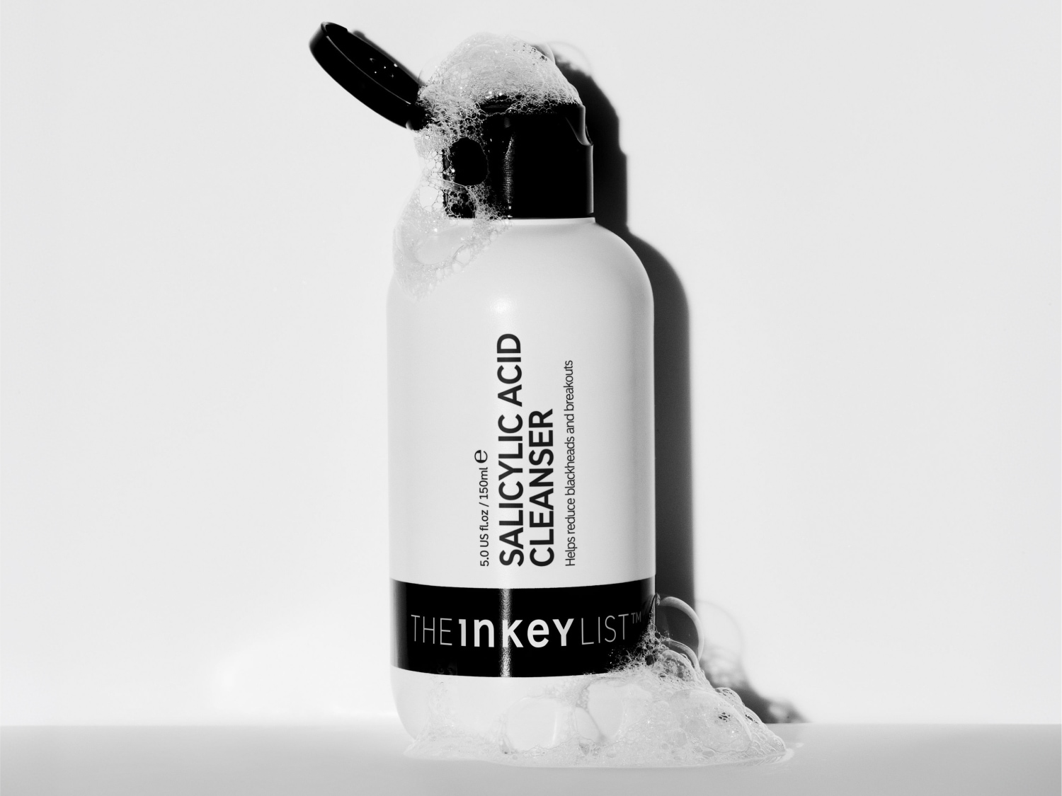 Eine weiße Flasche des Salicylic Acid Cleansers von The Inkey List mit geöffnetem schwarzem Klappverschluss steht auf weißem Untergrund, umgeben von Schaum.
