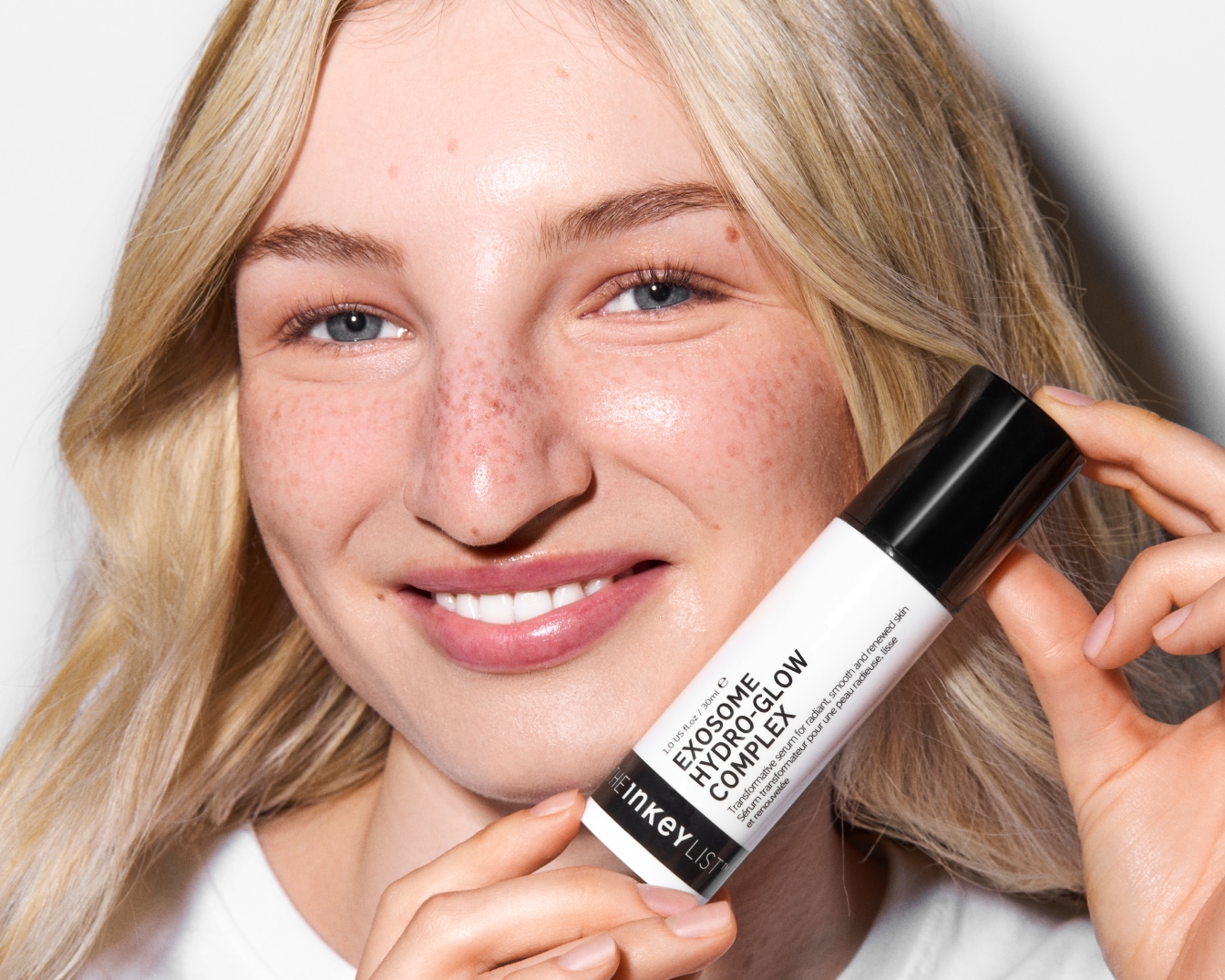 Eine lächelnde Frau mit blondem Haar und Sommersprossen hält ein weißes Fläschchen des Serums THE INKEY LIST EXOSOME HYDRO-GLOW COMPLEX in ihren Händen.