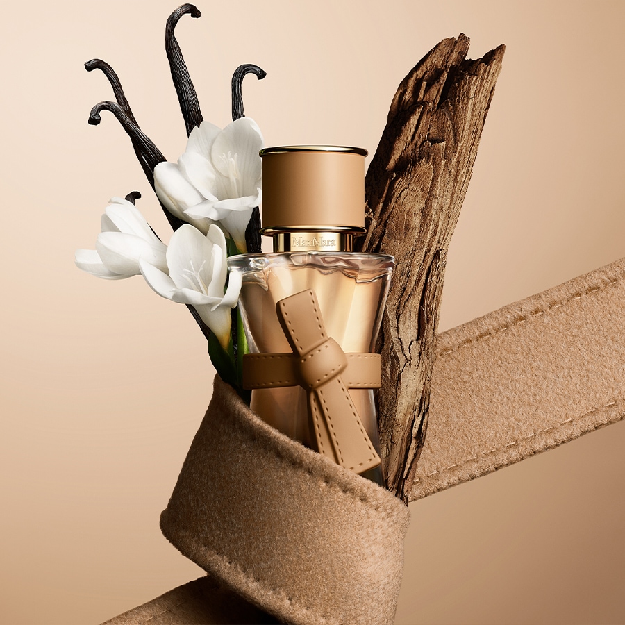 Ein Max Mara Parfumflakon mit beigefarbener Schleife und Kappe, umgeben von weißen Blüten, Vanilleschoten und einem Stück Holz, alles auf einem beigefarbenen Stoffband.