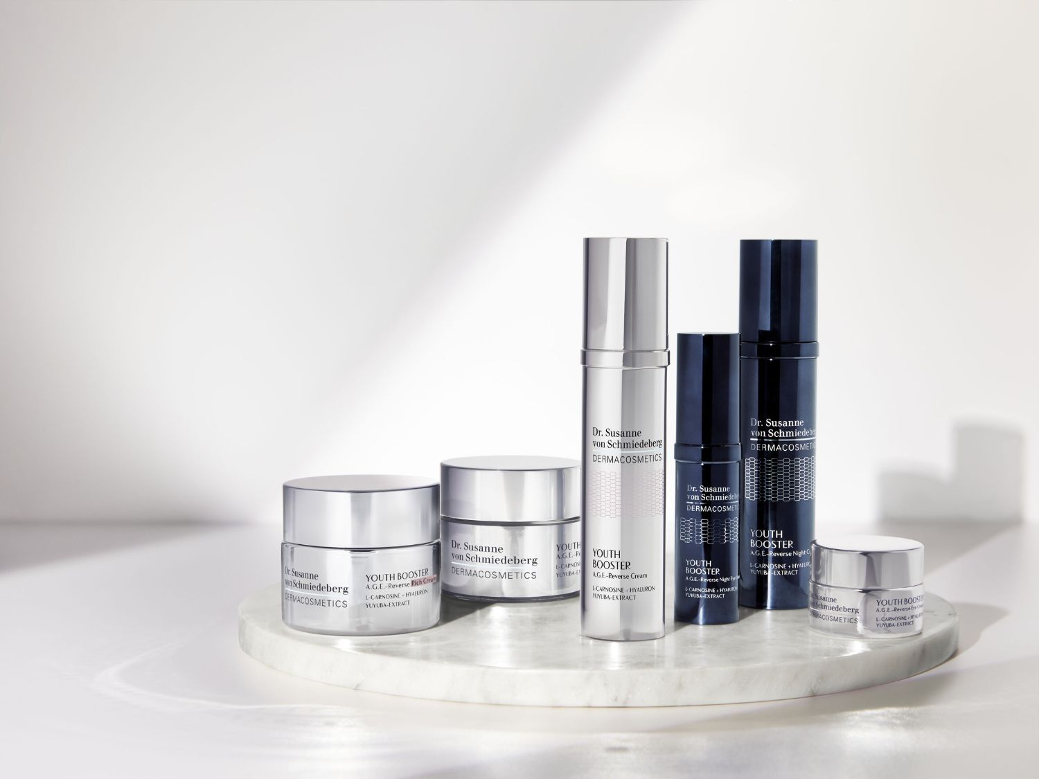 Šest produktů Dr. Susanne von Schmiedeberg DERMACOSMETICS Youth Booster, včetně krémů a sér v šedých a tmavě modrých obalech, je úhledně uspořádáno na bílém mramorovém podstavci.