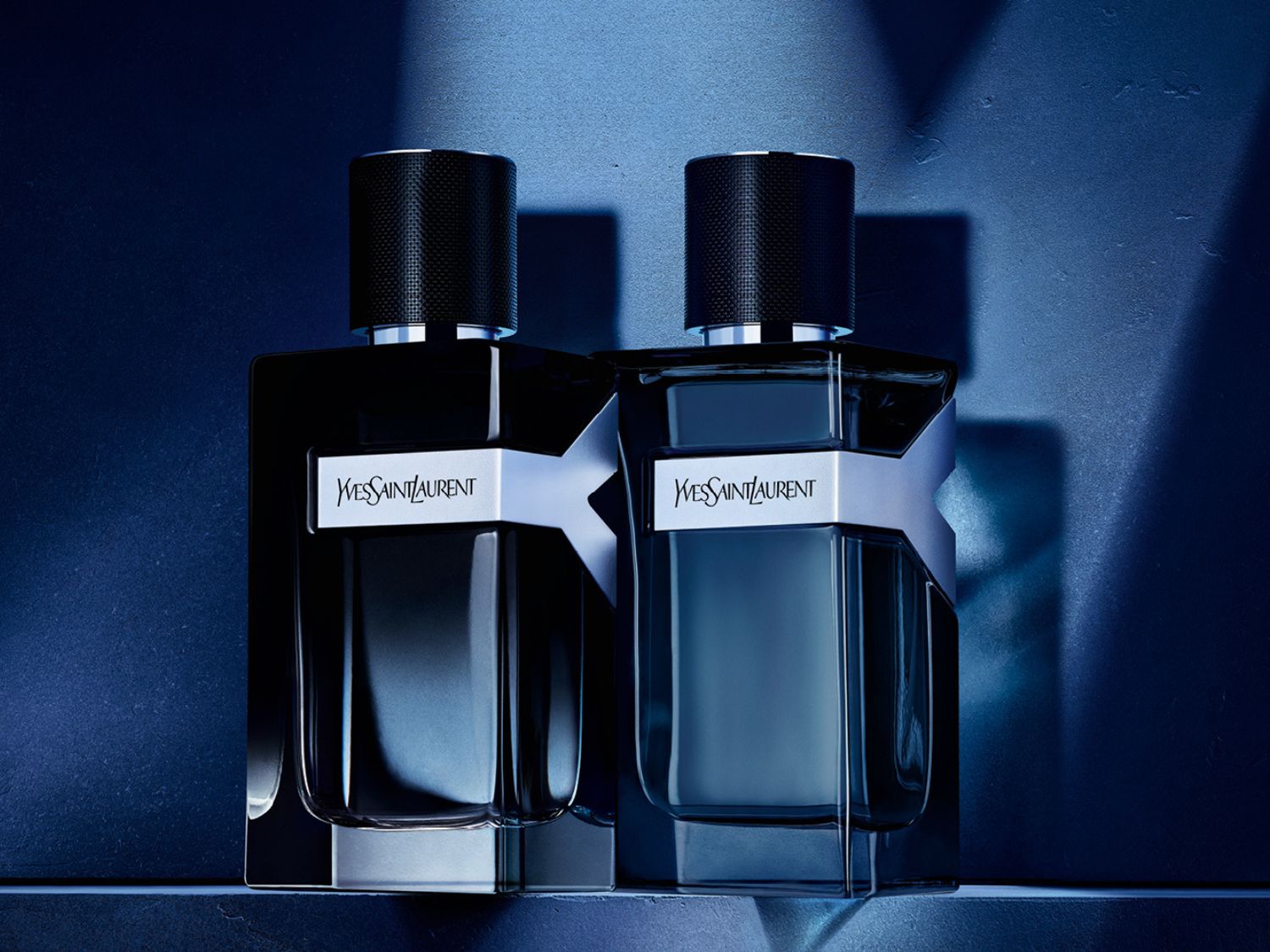Dvě lahvičky parfému Yves Saint Laurent Y Le Parfum s tmavě modrou tekutinou a stříbrným logem Y na tmavě modrém pozadí.