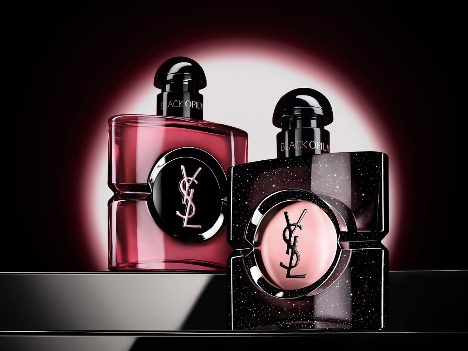 Dvě lahvičky parfému Black Opium od Yves Saint Laurent, jedna v průhledné růžové a druhá v černé se třpytkami, obě s logem YSL uprostřed, jsou umístěny na lesklém černém povrchu s červeným světlem v pozadí.