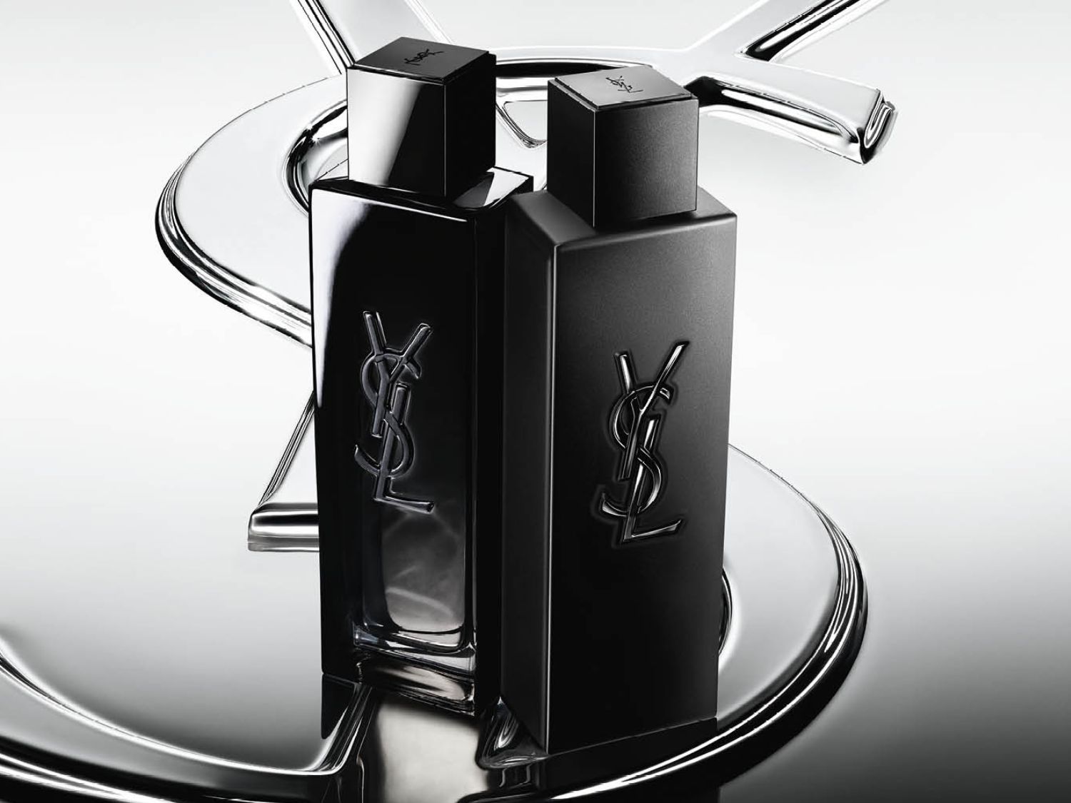 Dva elegantní černé flakony parfému Yves Saint Laurent s logem YSL jsou umístěny na lesklém stříbrném podkladu, který odráží jejich sofistikovaný design.