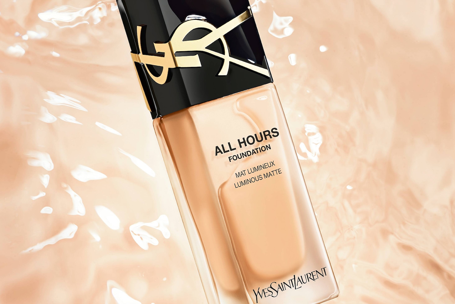 Podkład Yves Saint Laurent All Hours Foundation w beżowym odcieniu, z czarną nakrętką ozdobioną złotym logo YSL, leży na tle jasnobeżowej, falującej powierzchni.