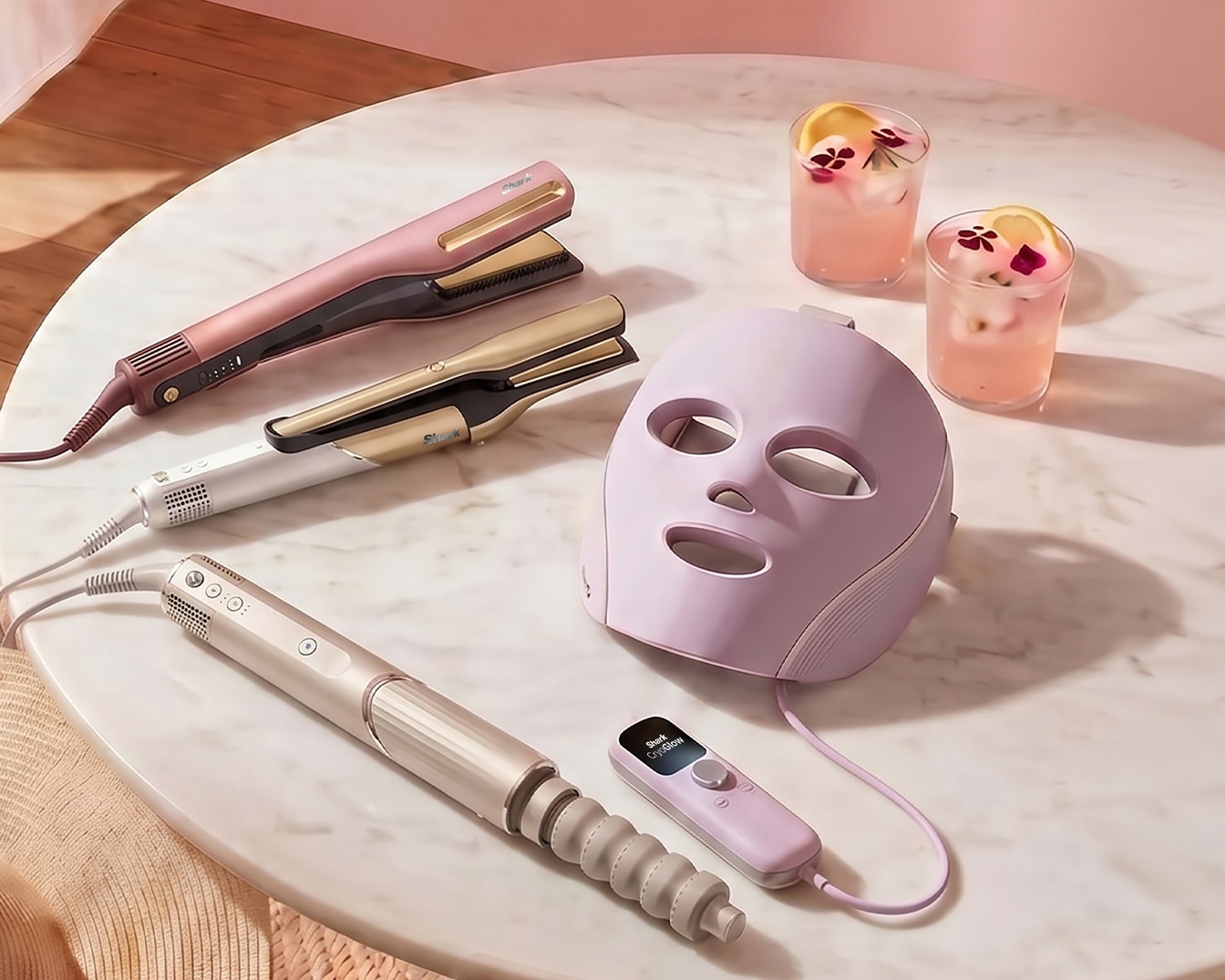 Découvrez une gamme d'outils de coiffure et de soins de la peau, comprenant des fers à lisser, un sèche-cheveux et un masque facial à LED, présentés sur une table en marbre avec deux verres de boisson rose.