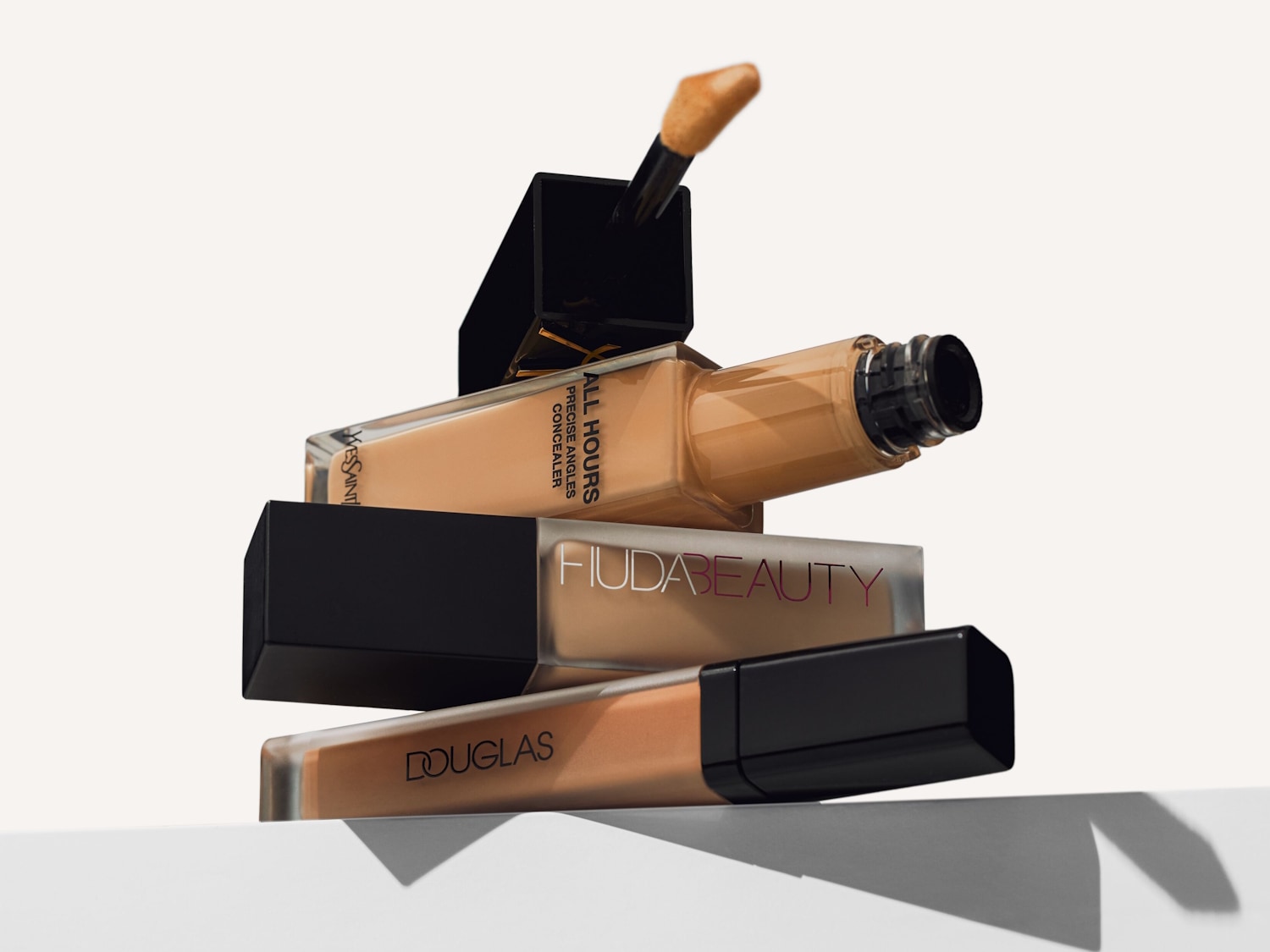 Tre flaconi di fondotinta e correttore di diverse tonalità, con il flacone Yves Saint Laurent in alto, Huda Beauty al centro e Douglas in basso, tutti con tappi neri e applicatori visibili.