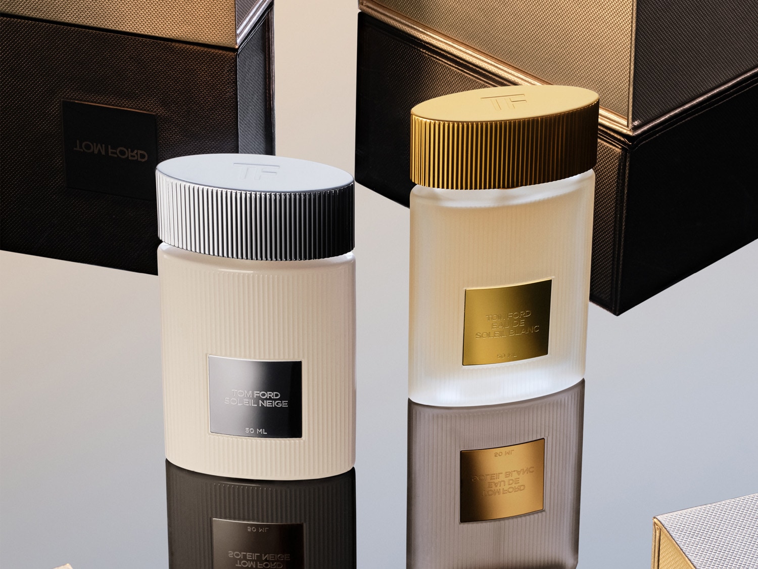 Zwei Tom Ford Parfümflaschen, Soleil Neige mit silbernem Deckel und Soleil Blanc mit goldenem Deckel, stehen umgeben von Geschenkboxen auf einem spiegelnden Untergrund.