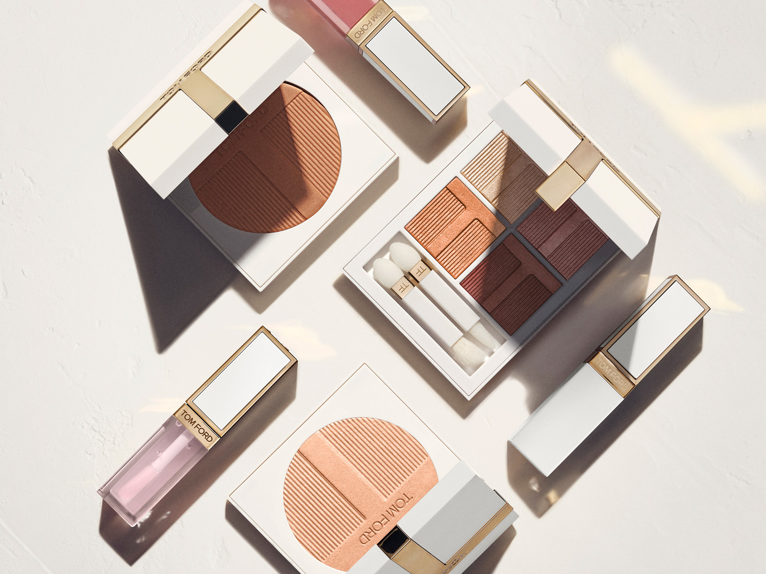 Eine Nahaufnahme von Tom Ford Beauty-Produkten, die elegant auf einer weißen Oberfläche angeordnet sind, darunter ein geöffneter Bronzer, eine Lidschattenpalette mit Pinseln und Lipglosse.