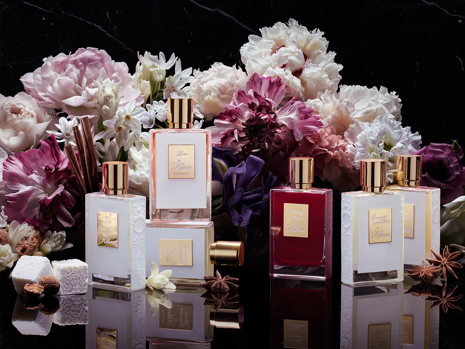 Eine Sammlung von Kilian Parfümflaschen, darunter "Rolling in Love" und "Good Girl Gone Bad", sind elegant auf einem glänzenden schwarzen Hintergrund arrangiert, umgeben von üppigen rosa, weißen und violetten Blüten.