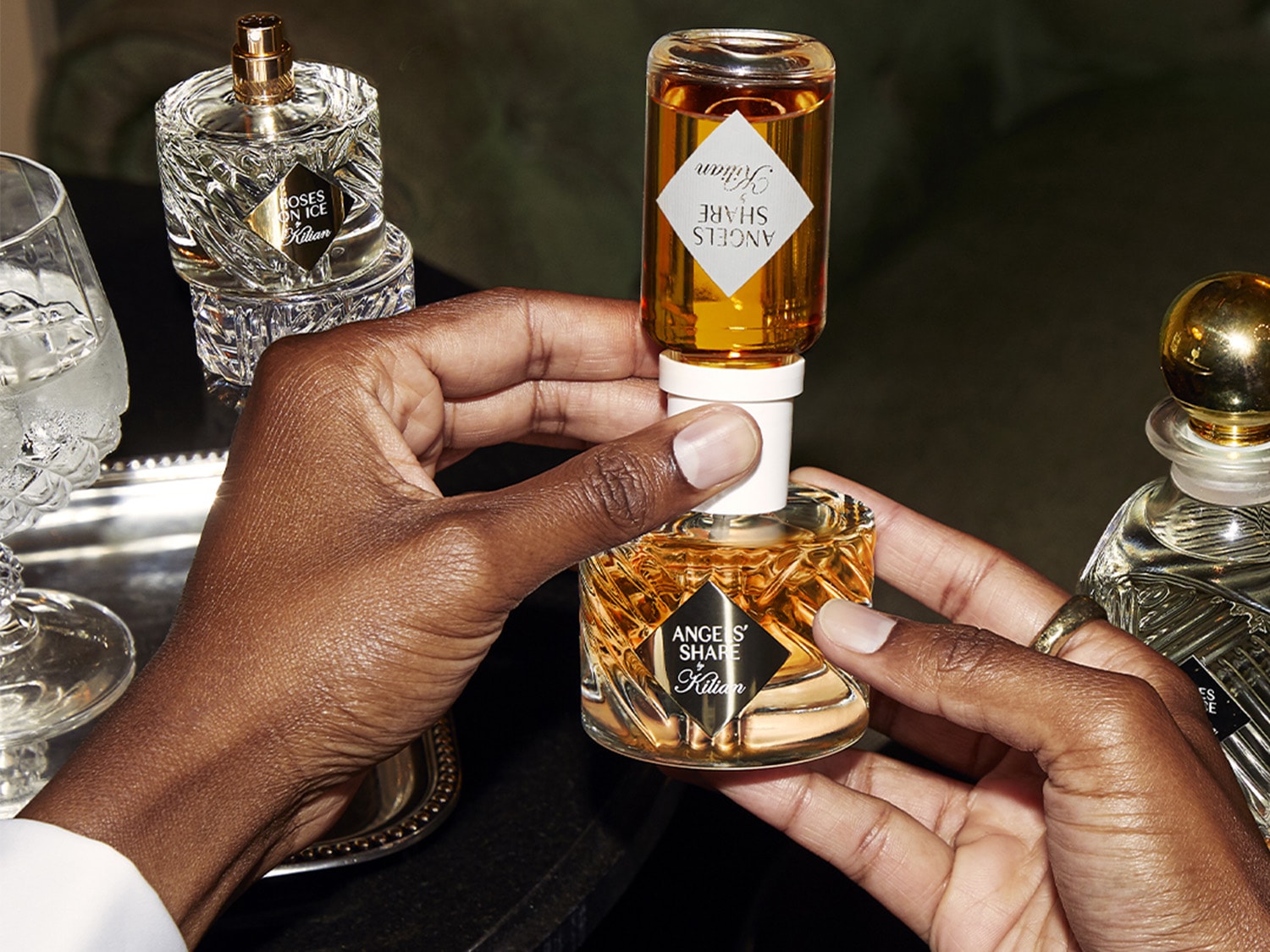 Zwei Hände halten einen goldenen Parfümflakon mit der Aufschrift „ANGELS SHARE by Kilian“ und füllen die Flüssigkeit mit einer Refill-Flasche ANGELS SHARE auf; im Hintergrund stehen ein weiterer Flakon und Kristallgläser.