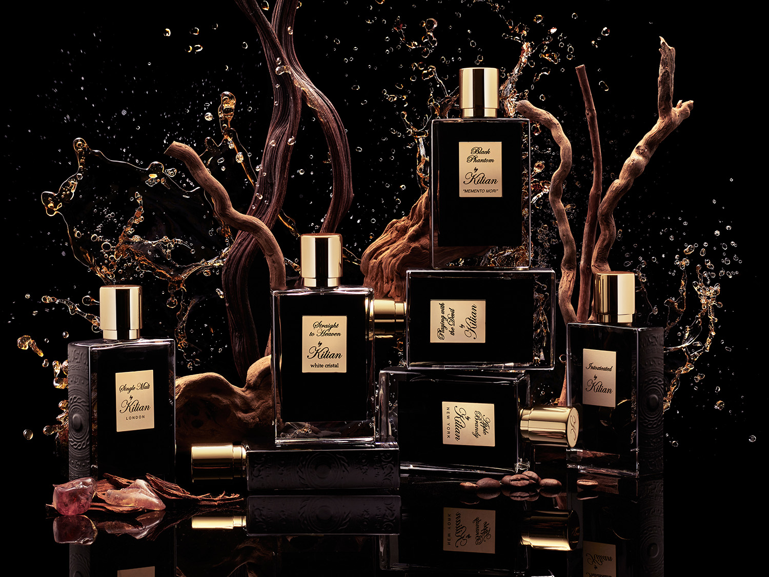 Eine Sammlung von Kilian-Parfümflaschen in Schwarz mit goldenen Etiketten und Kappen, umgeben von Holzstücken und spritzender Flüssigkeit auf schwarzem Hintergrund.