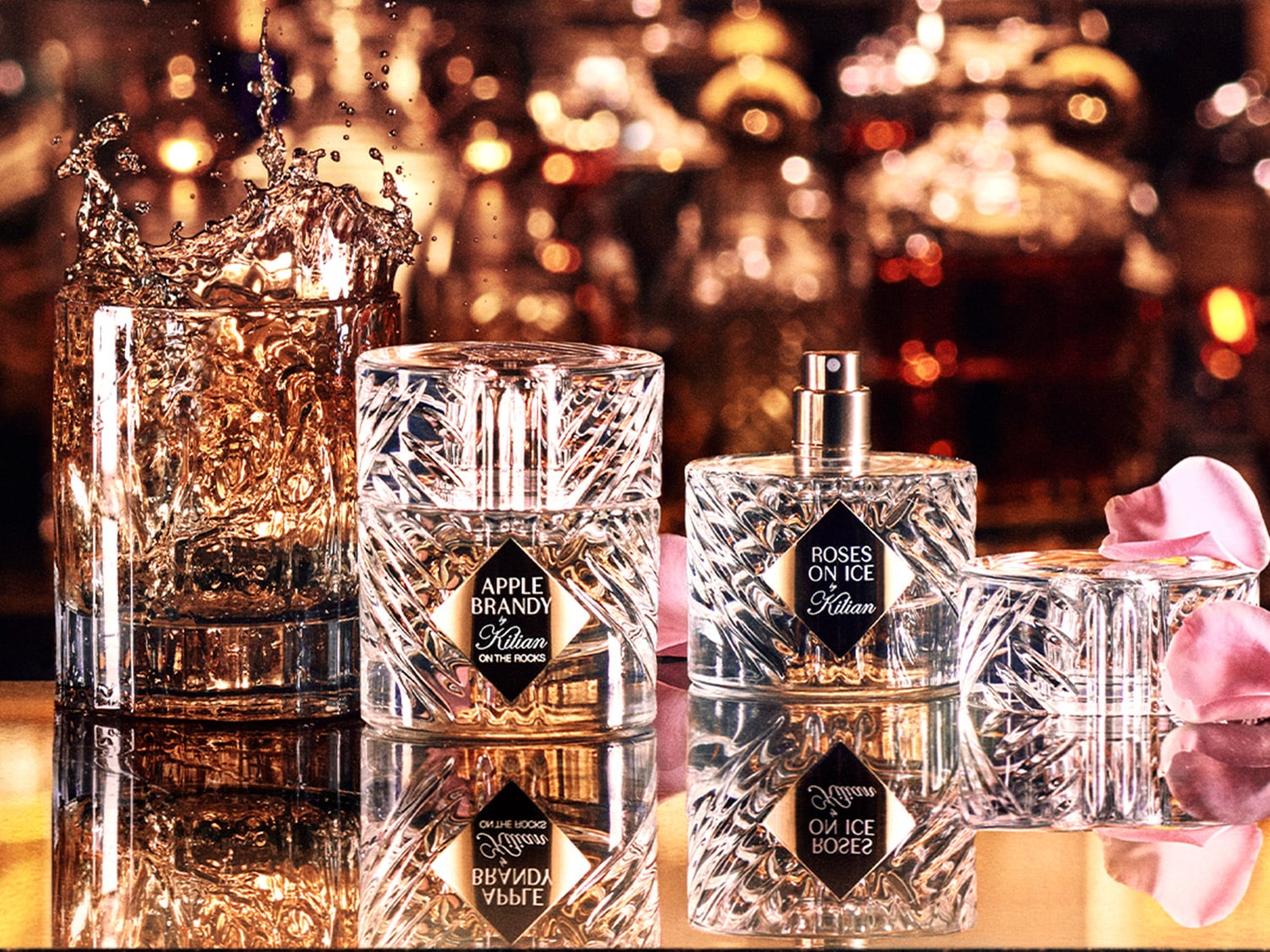 Zwei Kilian Parfümflaschen, "Apple Brandy" und "Roses on Ice", stehen auf einer spiegelnden Oberfläche neben einem Glas mit spritzender goldener Flüssigkeit und rosa Blütenblättern.