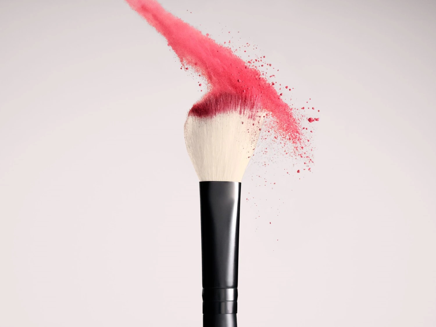 Un pennello da trucco con manico nero e setole chiare è cosparso di polvere rosa brillante che si diffonde nell'aria, creando un effetto dinamico e vivace.