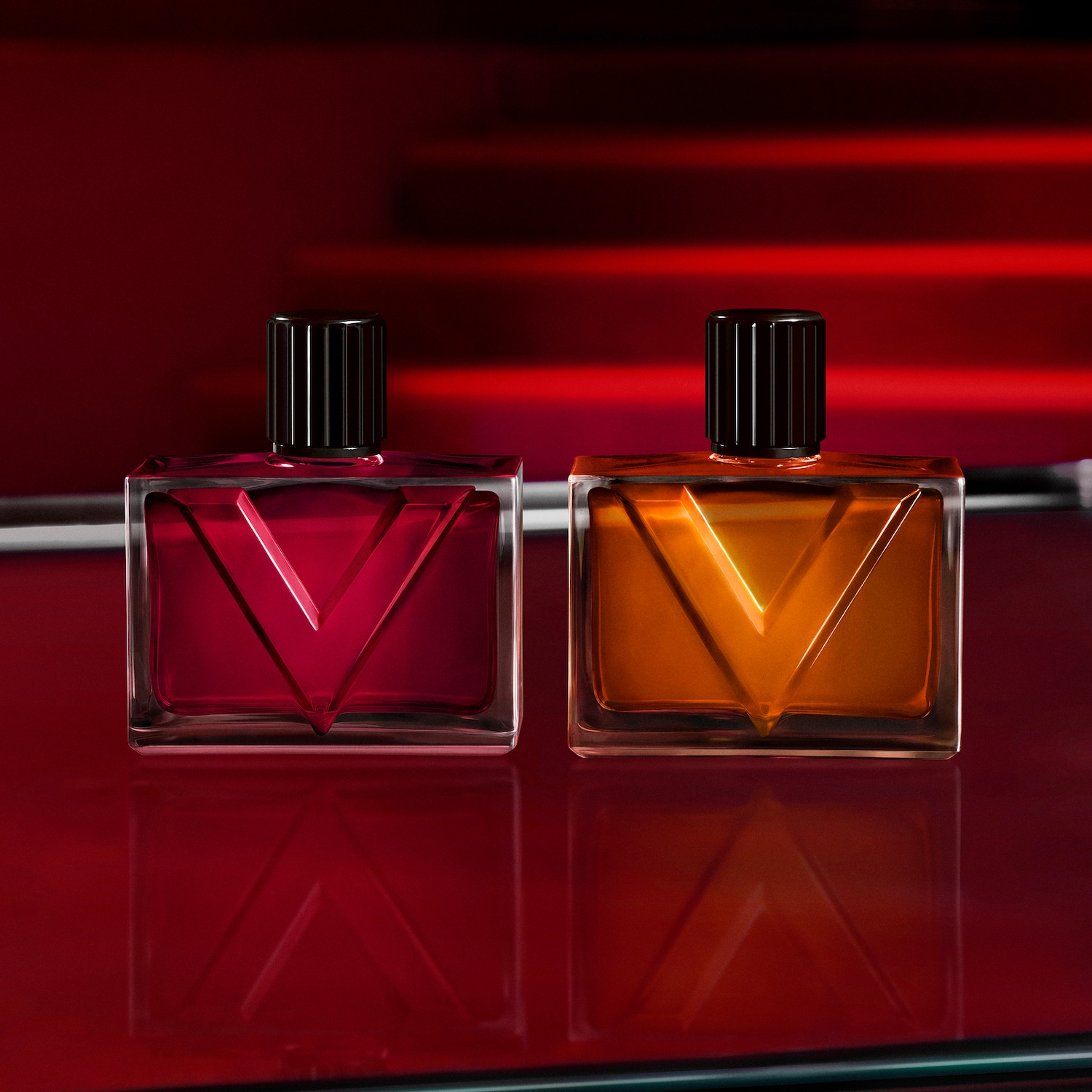 Zwei rechteckige Parfümflaschen stehen nebeneinander auf einer glänzenden roten Oberfläche, die linke Flasche ist rot und die rechte ist orange, beide haben schwarze, geriffelte Kappen und ein eingeprägtes V-förmiges Design auf der Vorderseite.