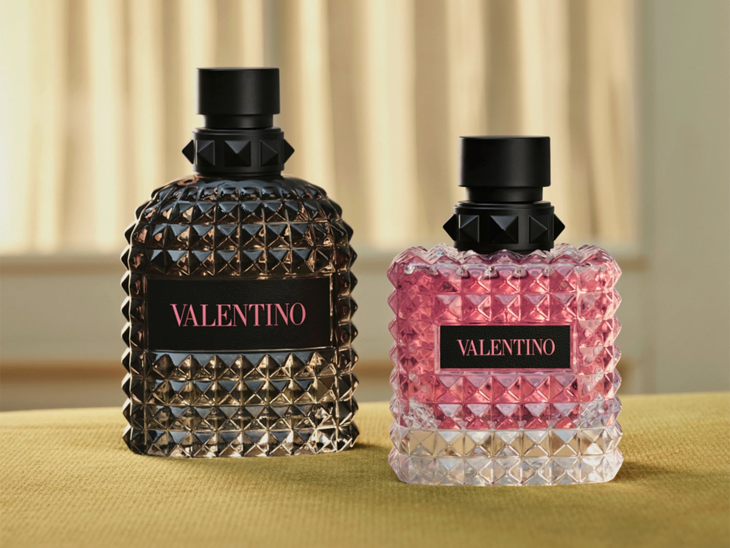Zwei Valentino Born in Roma Parfümflaschen stehen nebeneinander, eine in dunklem Glas mit schwarzem Etikett und pinker Schrift, die andere in klarem Glas mit pinkem Parfüm und schwarzem Etikett mit pinker Schrift, beide mit einem schwarzen, genieteten Verschluss.