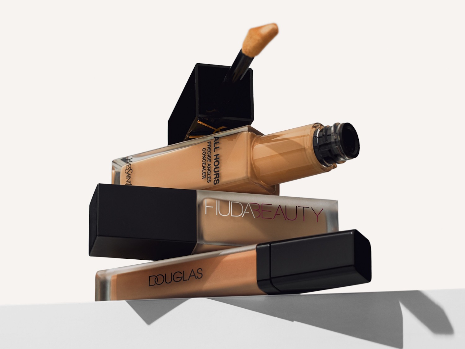 Tre flaconi di fondotinta e correttore di diverse tonalità, con il flacone Yves Saint Laurent in alto, Huda Beauty al centro e Douglas in basso, tutti con tappi neri e applicatori visibili.