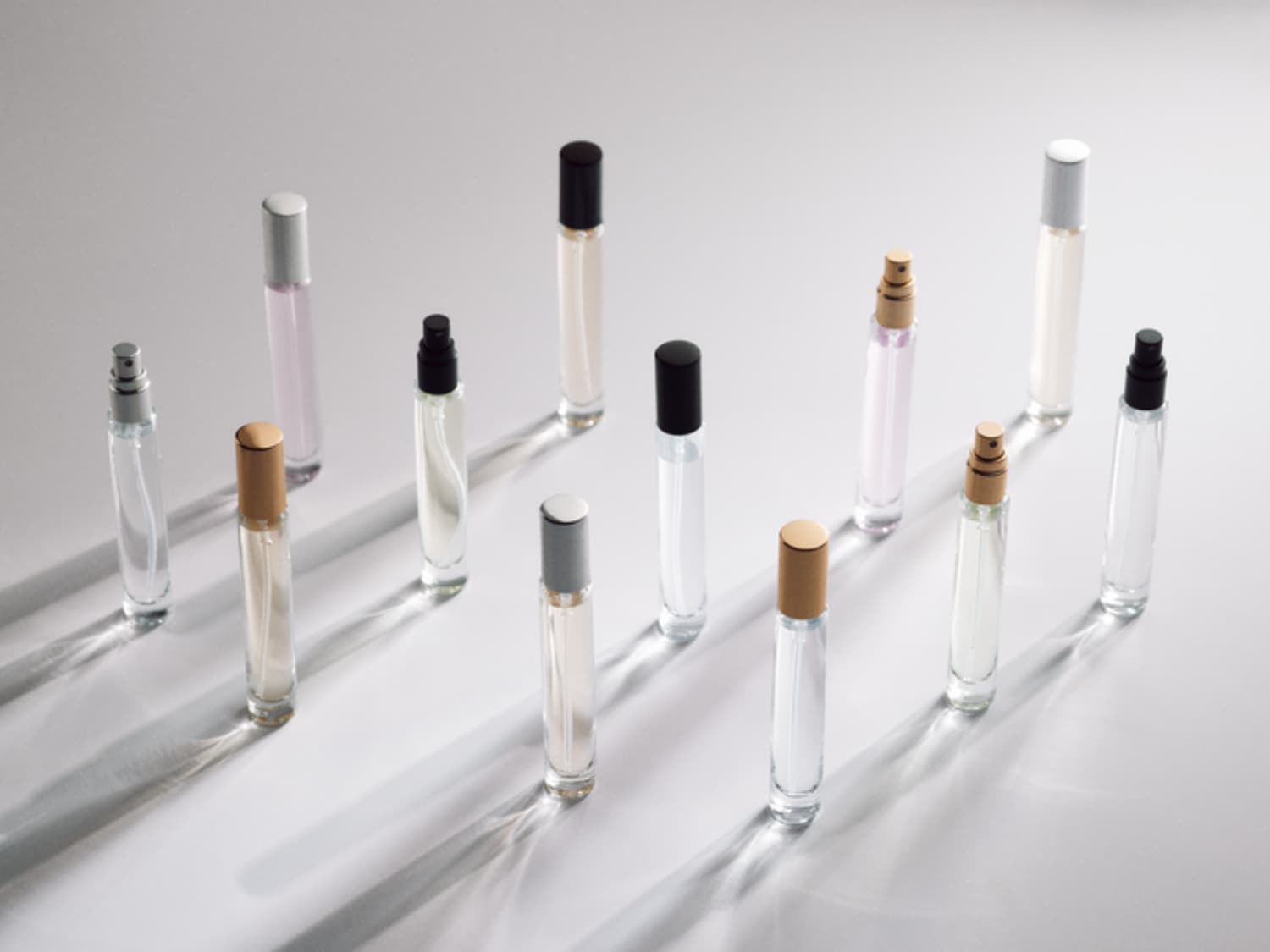 Een verzameling van elf slanke, glazen parfumflesjes met verschillende gekleurde doppen, variërend van zilver en goud tot zwart, staat op een witte ondergrond met lange schaduwen.
