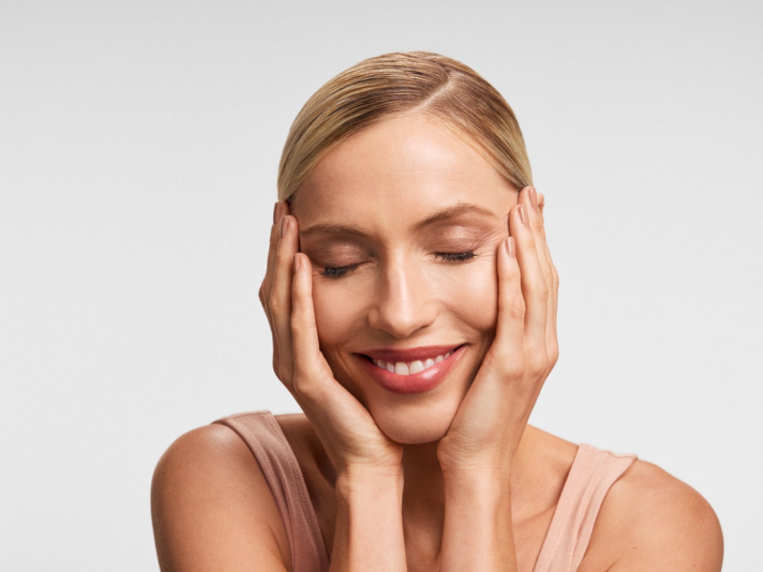 Een vrouw met blond haar en een perzikkleurige top glimlacht met gesloten ogen, haar handen zachtjes tegen haar wangen gedrukt, wat een gevoel van rust en tevredenheid uitstraalt.