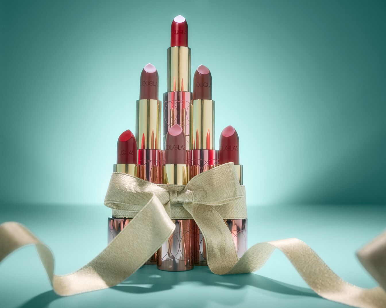 Un assortiment de rouges à lèvres Douglas, aux teintes rouges et roses, est présenté en forme de pyramide et orné d'un ruban doré, le tout sur un fond bleu-vert.
