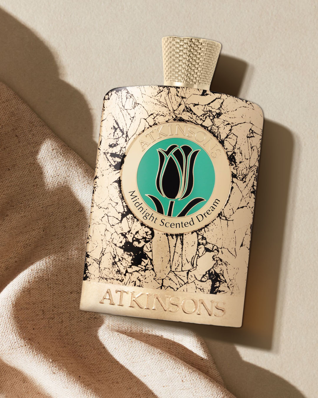 Perfumy Atkinsons Midnight Scented Dream w beżowym flakonie ze złotymi i czarnymi pęknięciami, z okrągłą etykietą z czarnym tulipanem na turkusowym tle, leżące na beżowym materiale.