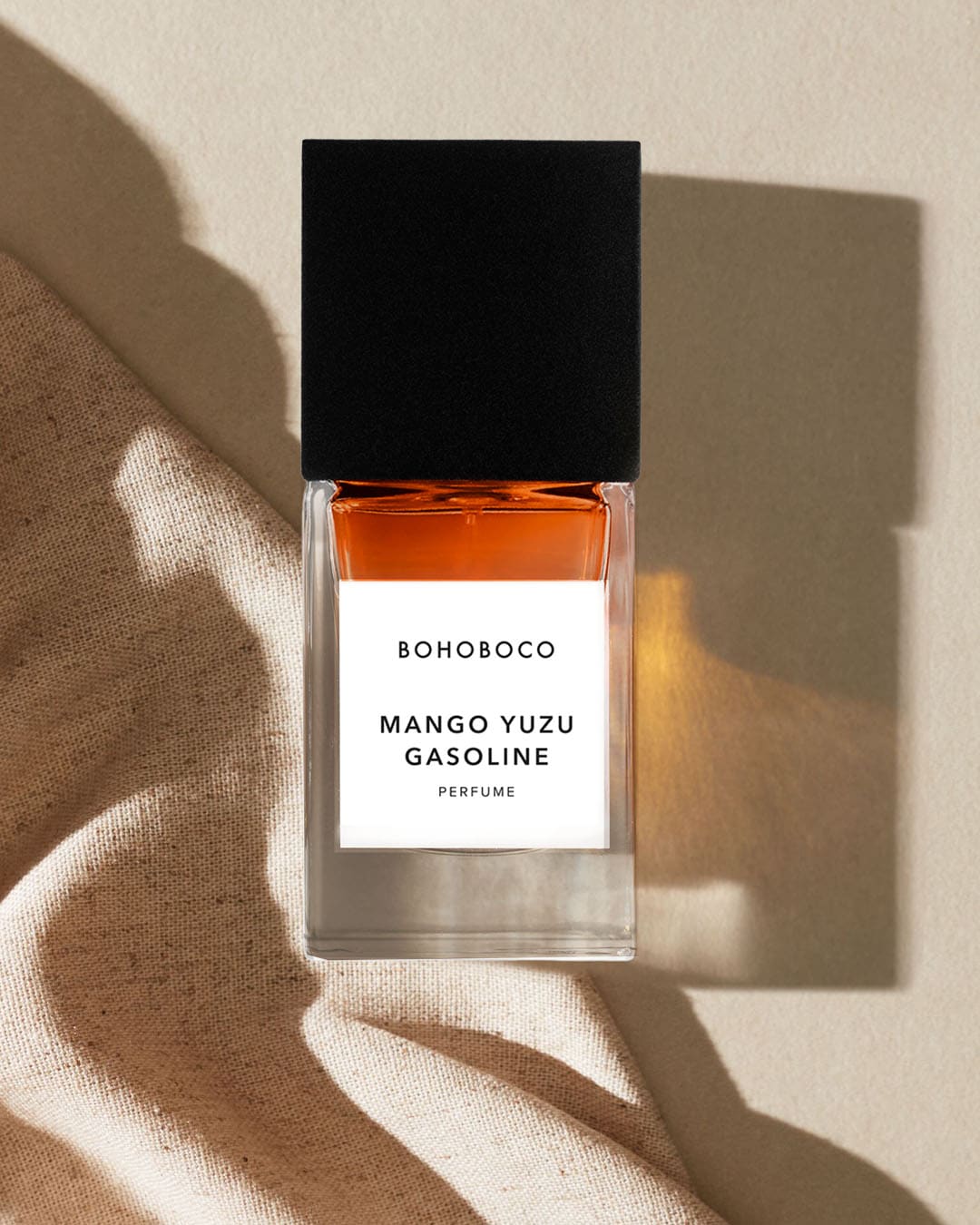 Perfumy BOHOBOCO Mango Yuzu Gasoline w prostokątnej butelce z czarną zakrętką, z bursztynowym płynem, stojące na beżowym tle z jasnobrązową tkaniną.