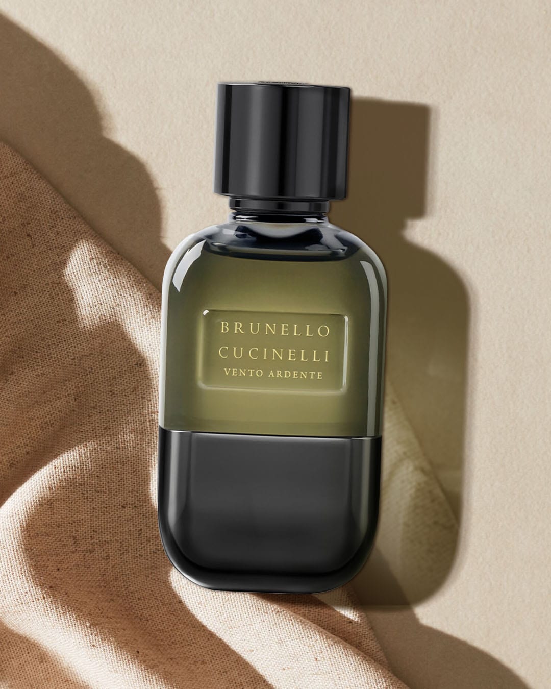Brunello Cucinelli Vento Ardente to perfumy w eleganckiej butelce z ciemnozielonego szkła z czarnym korkiem, leżące na beżowym materiale, idealne dla osób ceniących luksus i wyrafinowanie.