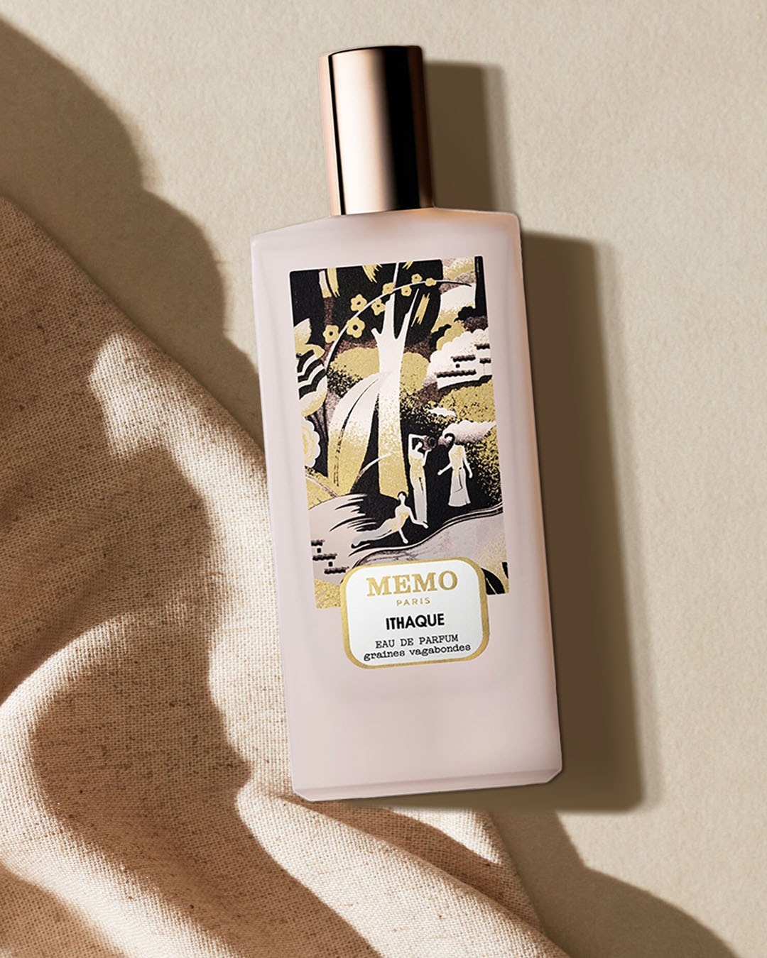 Flakon perfum Memo Paris Ithaque Eau de Parfum o pojemności 75 ml, z matowego szkła w kolorze jasnoróżowym, z miedzianą nakrętką i etykietą z czarno-złotym wzorem przedstawiającym drzewa i postacie, leży na beżowym materiale.