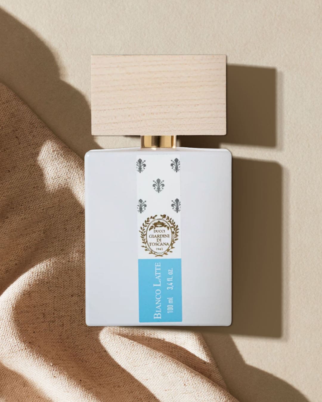 Biała butelka perfum Bianco Latte o pojemności 100 ml z drewnianą nakrętką i etykietą z logo Ducci Giardini di Toscana, leżąca na beżowym tle z brązową tkaniną.
