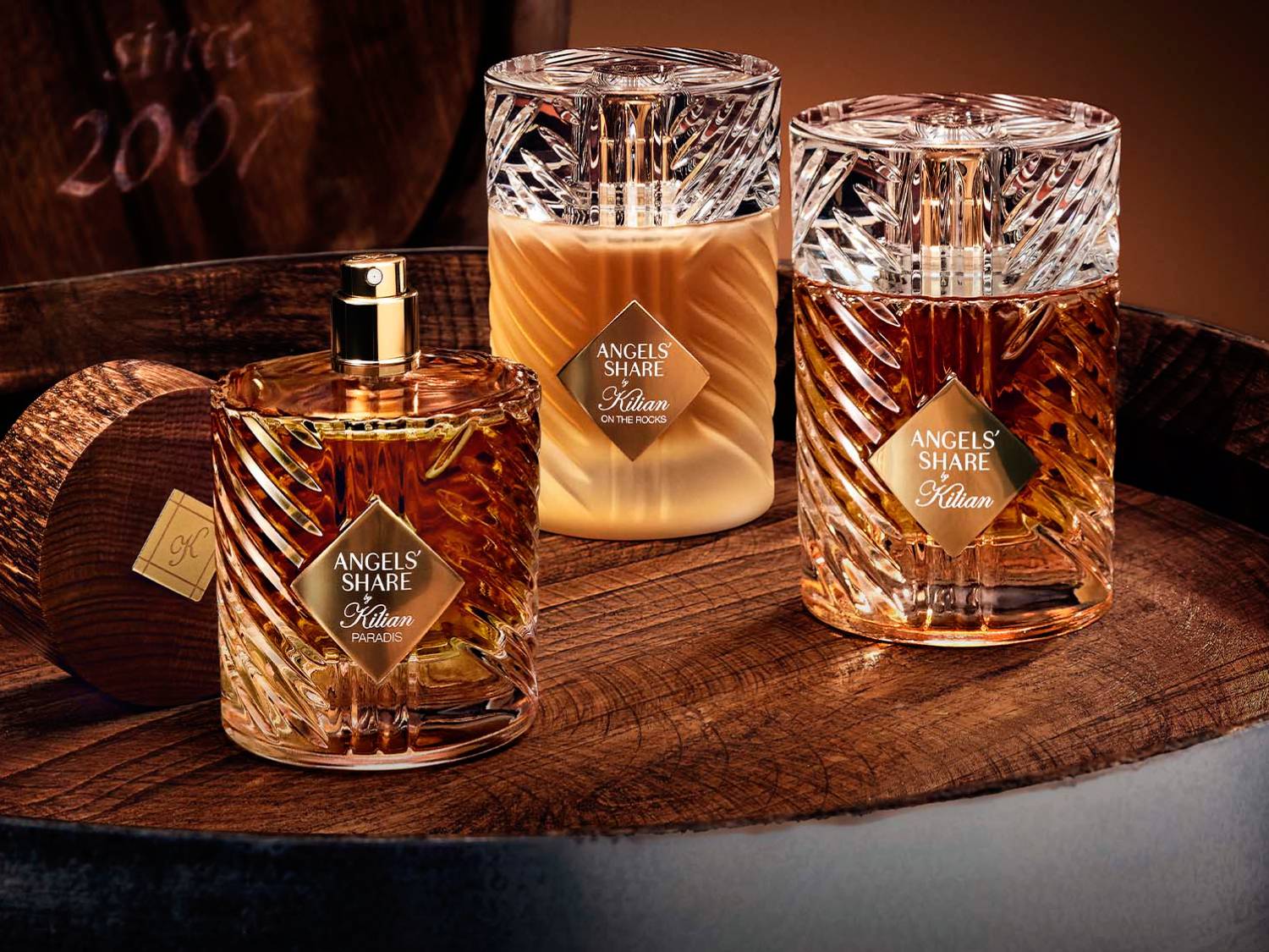 Trzy butelki perfum Kilian Angels' Share, dwie z bursztynowym płynem i jedna z kremowym, stoją na drewnianej beczce, a na tle widnieje napis „since 2007”.